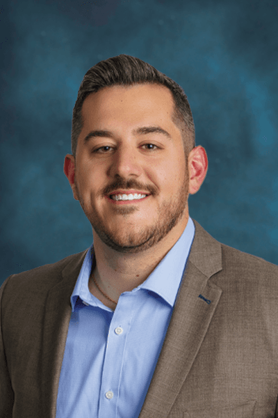 Smiling man with dark hair and beard in a tan blazer and blue shirt