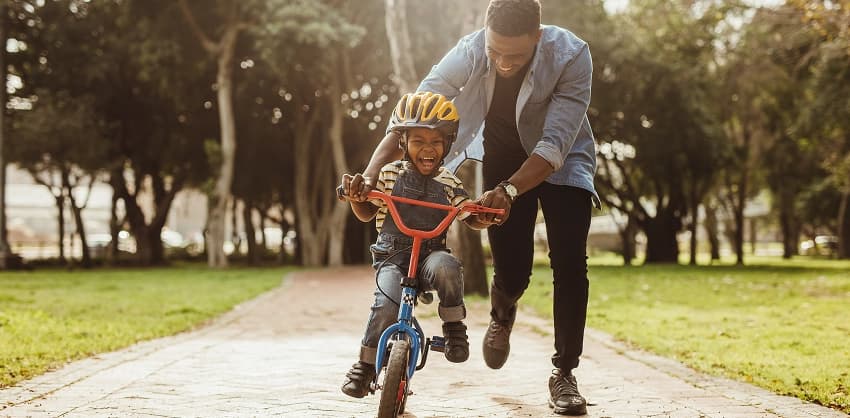 A father helping his laughing child learn to ride a bike in a sunny park.