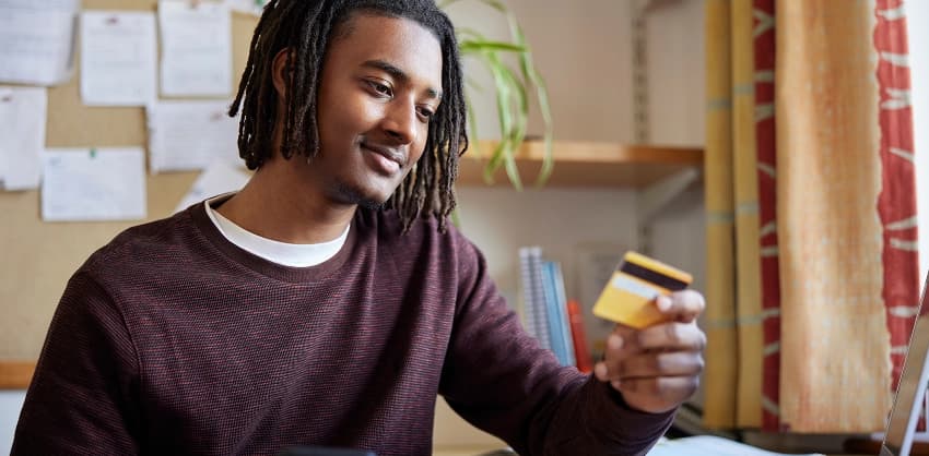 Young man holding a credit card while browsing online banking on a laptop at a desk