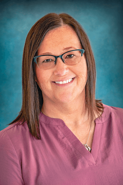 Smiling female staff member with brown hair and glasses wearing a mauve medical scrub top