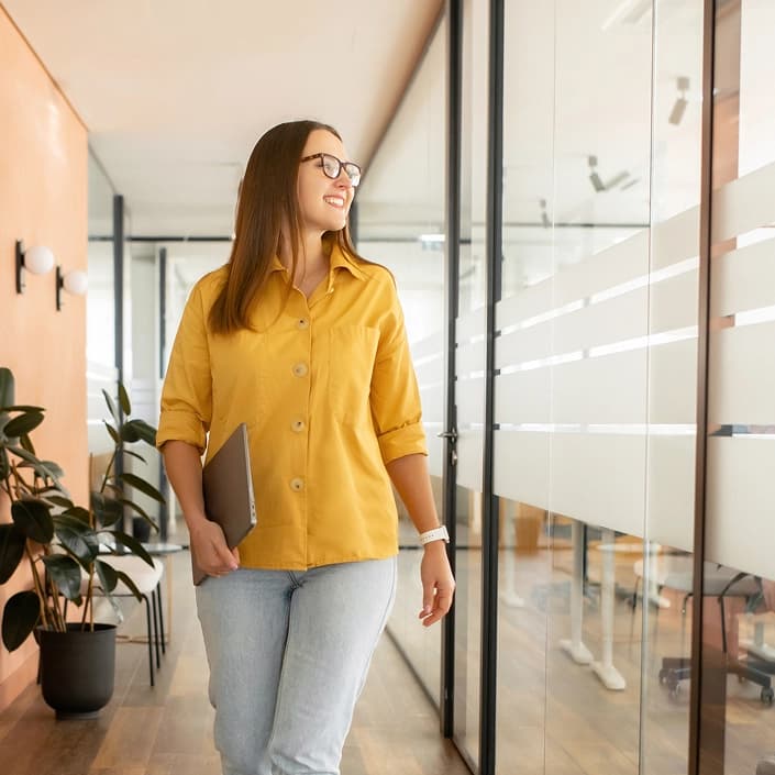 Smiling woman in yellow shirt carrying a laptop while walking through a modern office hallway