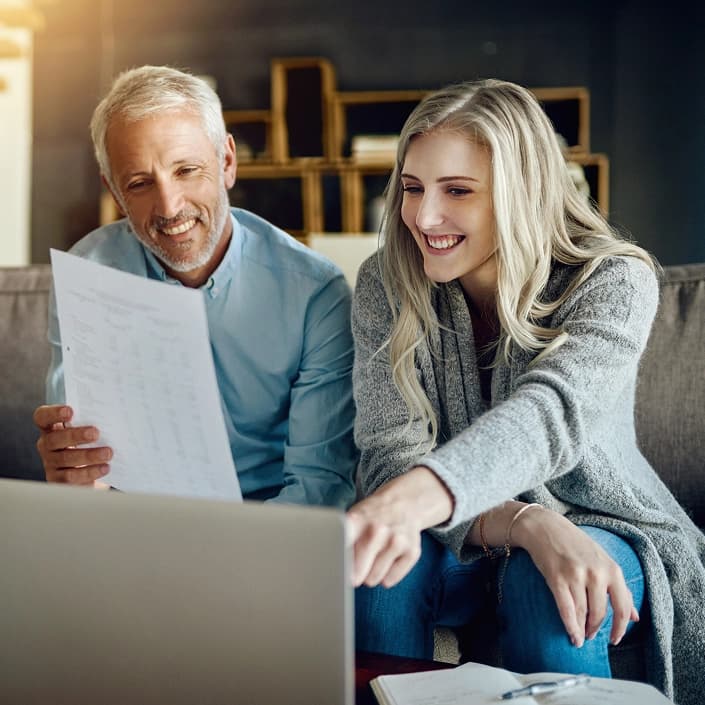 Two people smiling while reviewing financial documents on a laptop at home