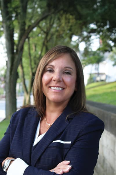 Smiling woman with dark hair in a navy blazer, arms crossed, posing outdoors