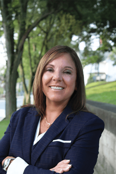 Smiling woman with dark hair in a navy blazer, arms crossed, posing outdoors