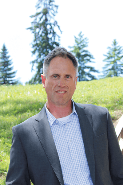 Man with dark hair wearing a gray blazer and plaid shirt outdoors on a grassy hill
