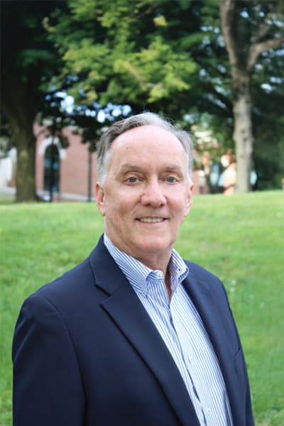 Man with gray hair wearing a navy blazer and striped shirt smiling outdoors