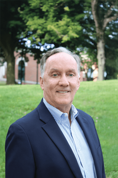 Man with gray hair wearing a navy blazer and striped shirt smiling outdoors