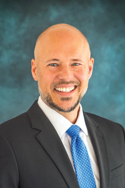 Smiling bald man in a dark suit and blue tie against a teal background