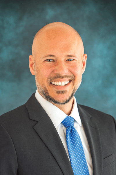 Smiling bald man in a dark suit and blue tie against a teal background