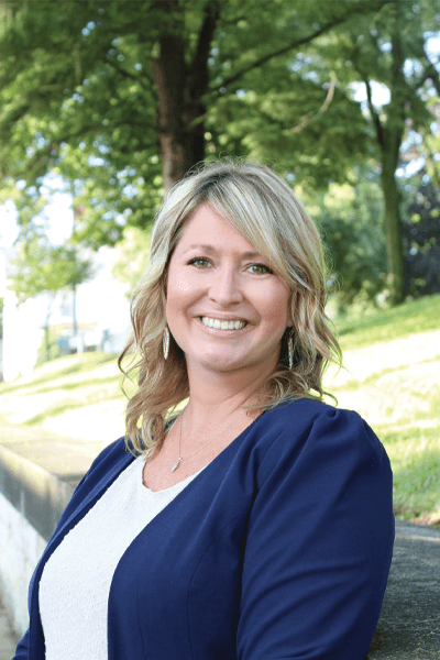 Smiling woman with blonde hair wearing a navy blazer outdoors among trees