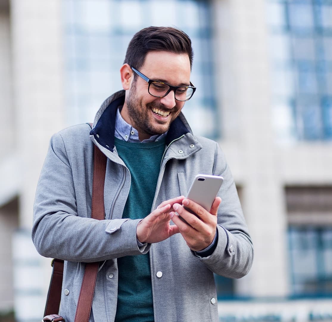 Smiling man with glasses using a smartphone outdoors near a modern building
