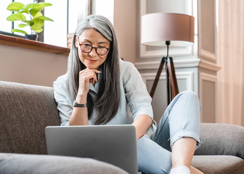 Woman with gray hair relaxing on a sofa, browsing online banking on a laptop