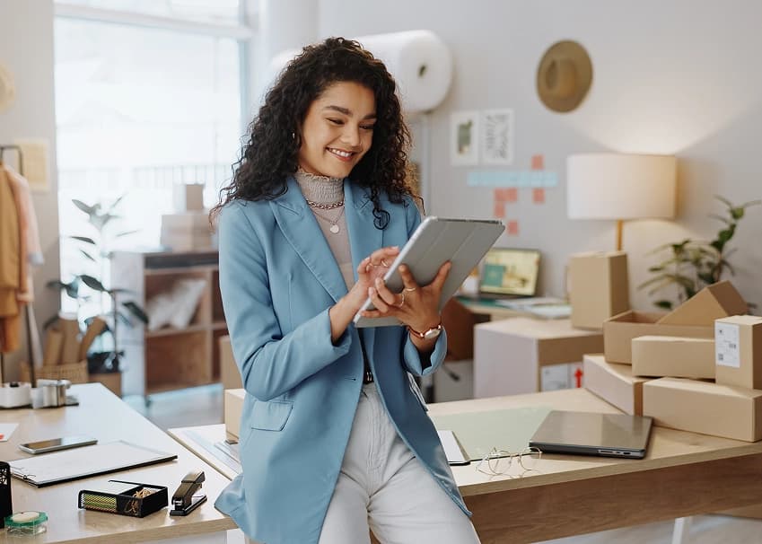 Smiling woman in a blue blazer using a tablet in a retail business workspace with shipping boxes