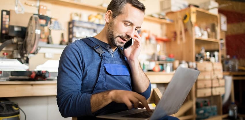 A man in a workshop apron talking on the phone while reviewing documents on a laptop