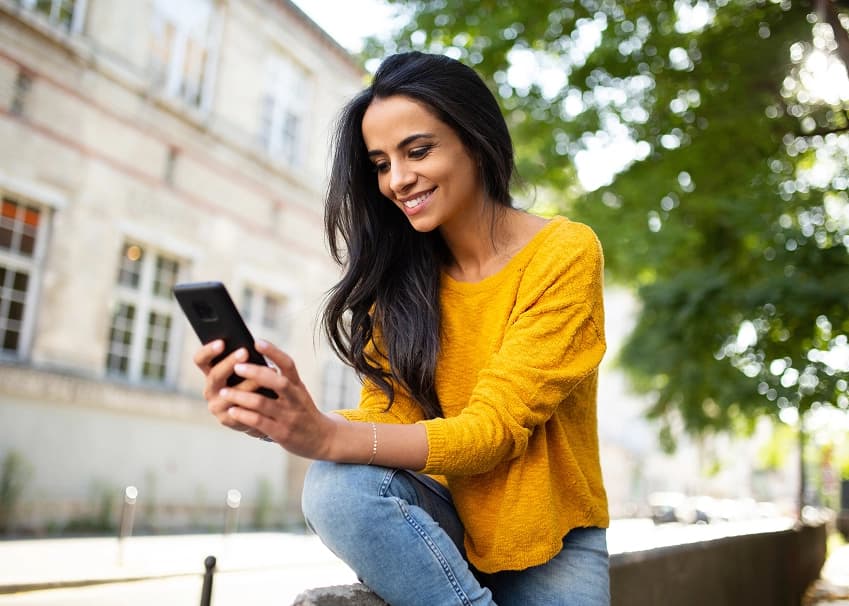 Woman in yellow sweater smiling while using her smartphone outdoors