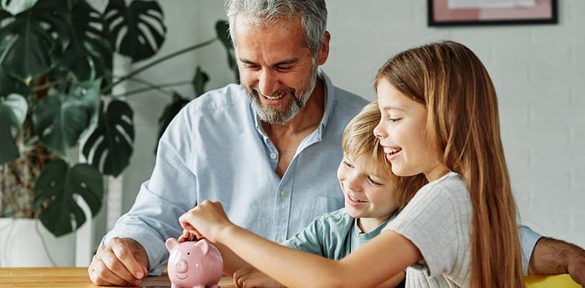 A father and two children smiling while putting coins into a pink piggy bank.