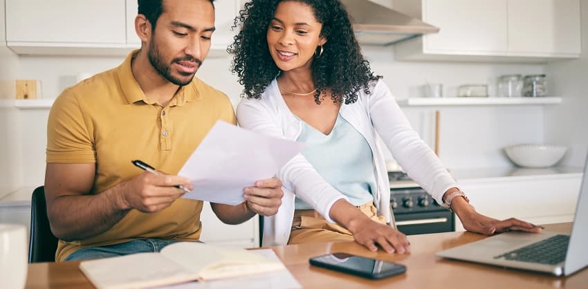 Couple reviewing mortgage loan documents together at a kitchen table with a laptop and smartphone nearby