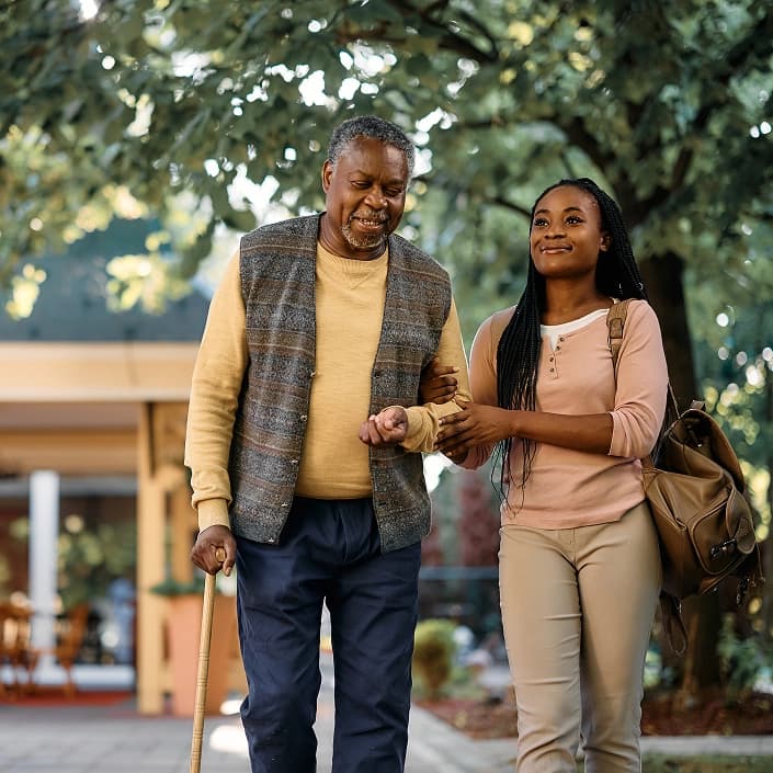 A young woman helping a man with a cane walk outdoors, symbolizing personal care and support.