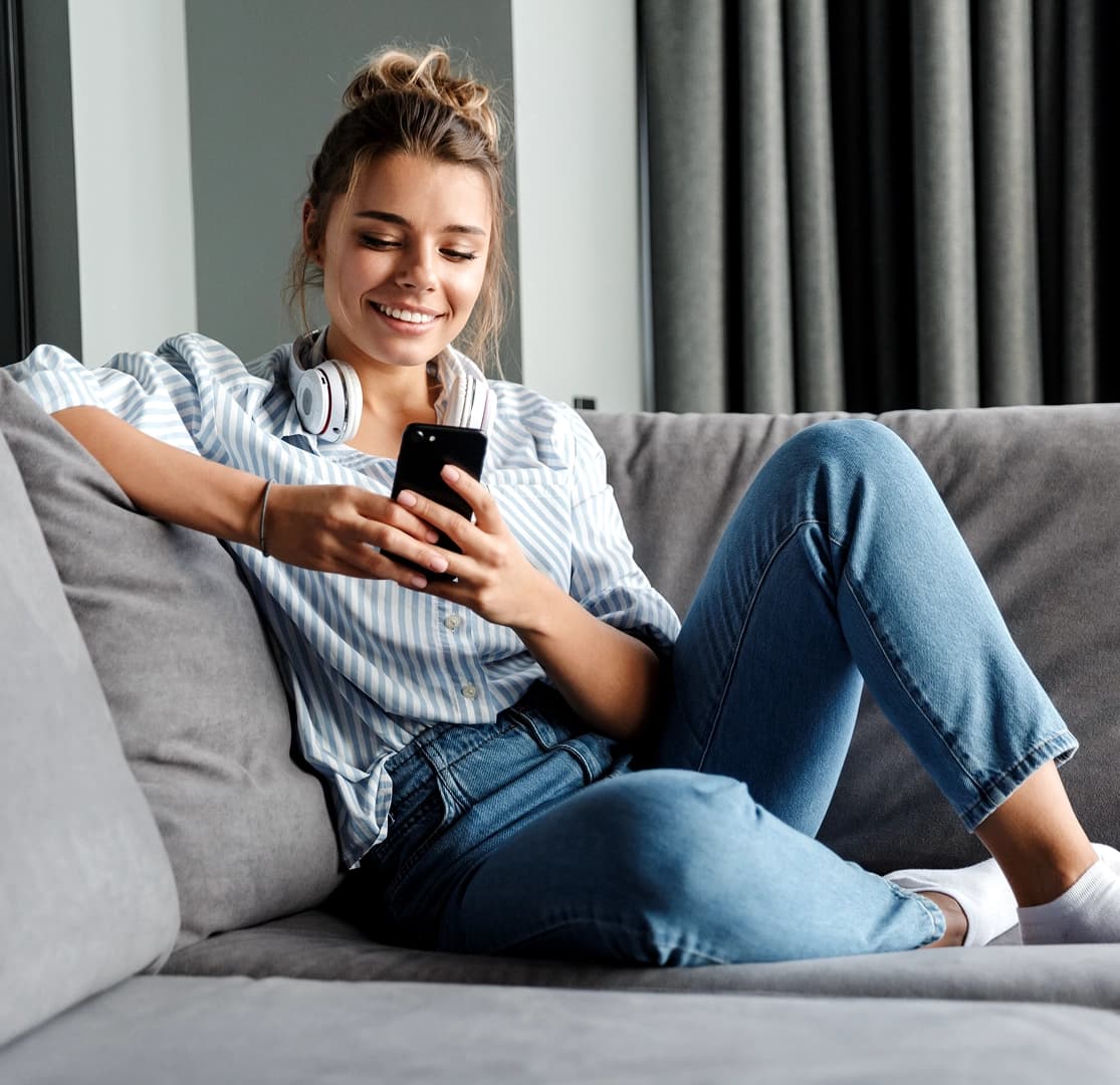 Young woman smiling while using her smartphone on a cozy gray couch with headphones around her neck