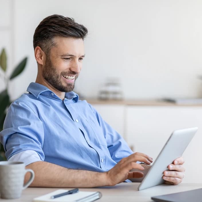 Smiling man in a blue shirt using a tablet at a desk with coffee nearby