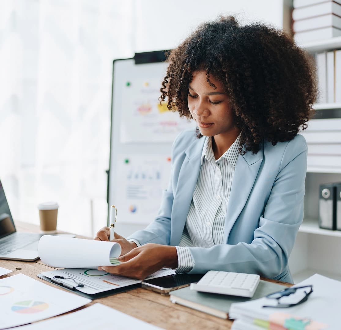 Woman in a light blue blazer reviewing financial documents at a desk with charts and a calculator
