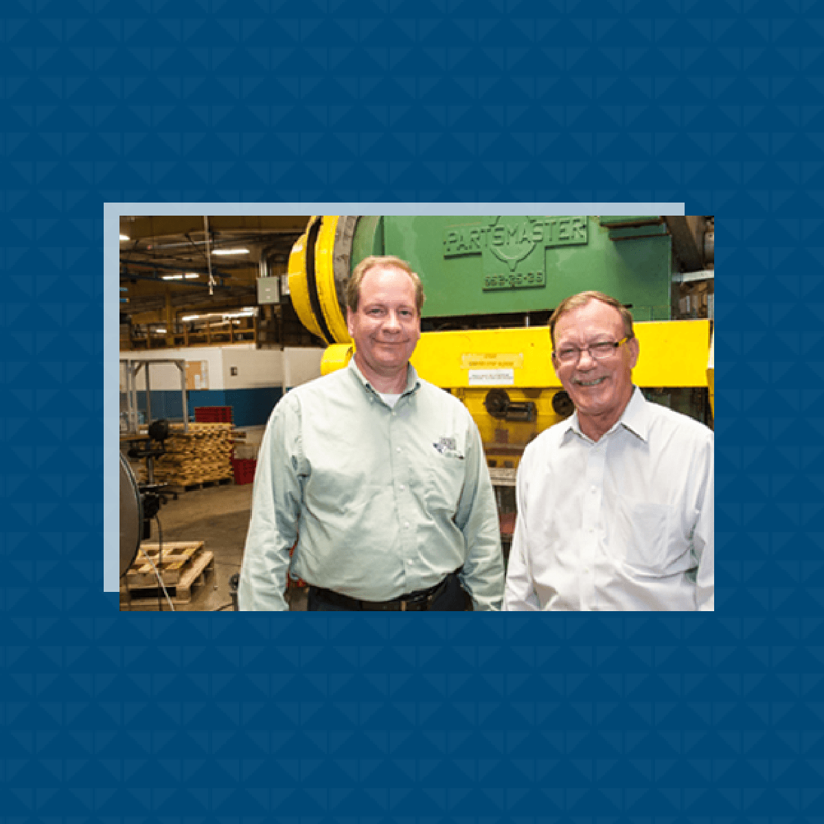 Two men smiling in a manufacturing facility beside a Partsmaster press machine.