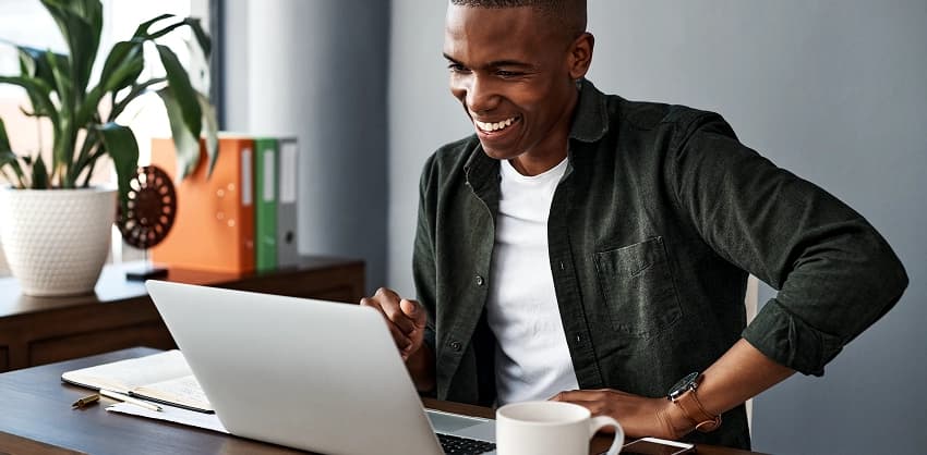 Smiling man viewing his laptop screen at a home office desk with coffee