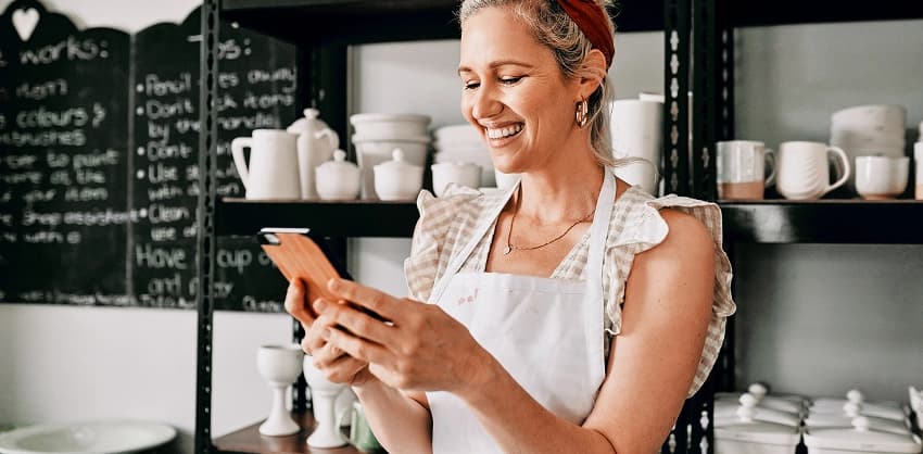 Woman in apron smiling while using a smartphone in a pottery shop