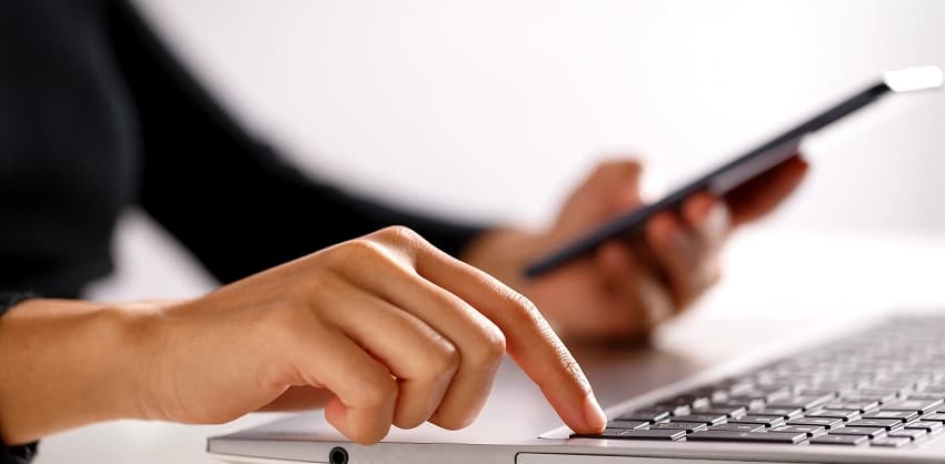 Person using a laptop trackpad while holding a smartphone, managing online banking tasks
