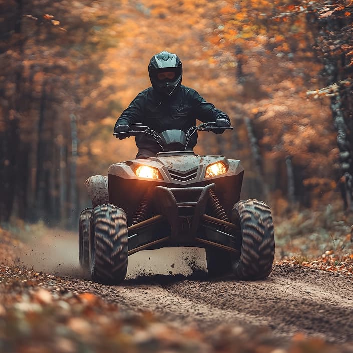 Rider in full helmet and gear driving a muddy ATV along a dirt trail through autumn woods