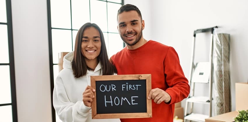 Couple smiling and holding a chalkboard sign that reads "Our First Home"