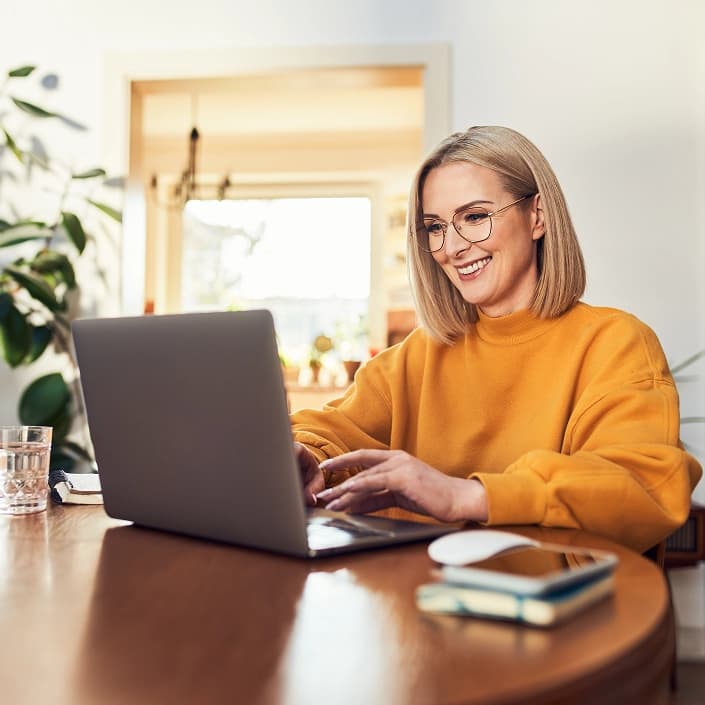 A smiling woman in a yellow sweater using a laptop at home for financial planning