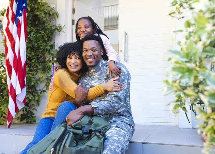 Smiling military serviceman in uniform embracing his family on their home's front steps with an American flag nearby