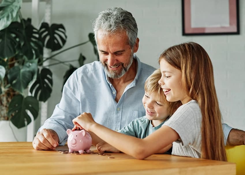 A man and two children happily putting coins into a pink piggy bank at a wooden table