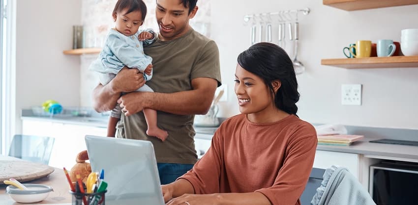 Young family in a kitchen, woman smiling at laptop while man holds a baby, managing online banking.