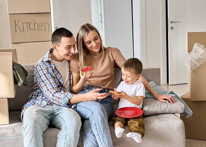 Young family relaxing on a couch surrounded by moving boxes, sharing a snack in their new home