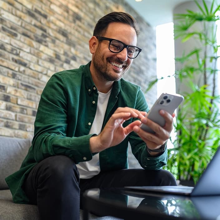 Smiling man in glasses using a smartphone for online banking while sitting on a couch