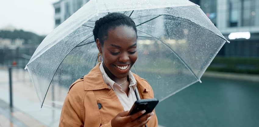 Smiling woman checking her phone while holding a clear umbrella in the rain