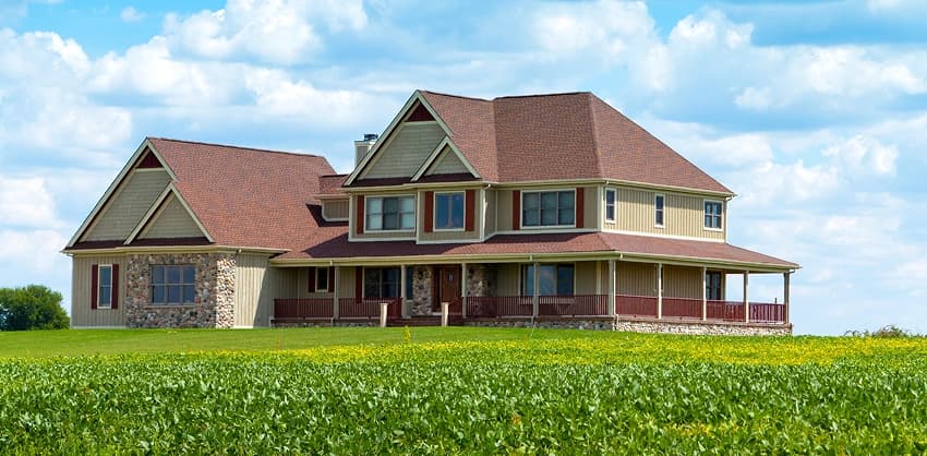 Large two-story farmhouse with a wraparound porch surrounded by green fields under a blue sky