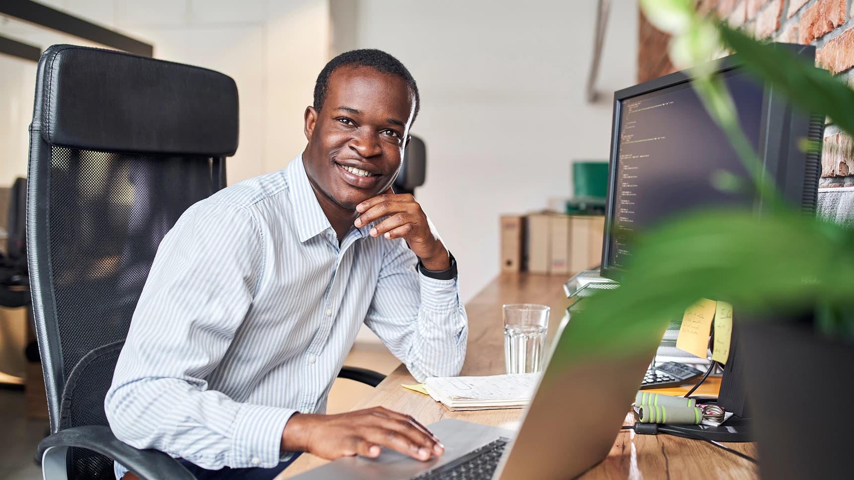 Man smiling at his desk with a laptop and monitor displaying code in a modern office