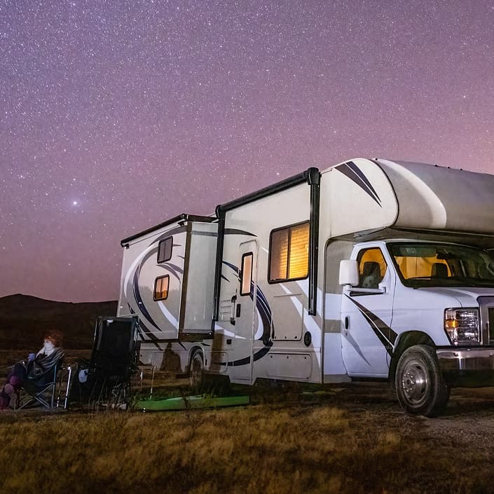 Person relaxing outside a lit RV under a starry night sky