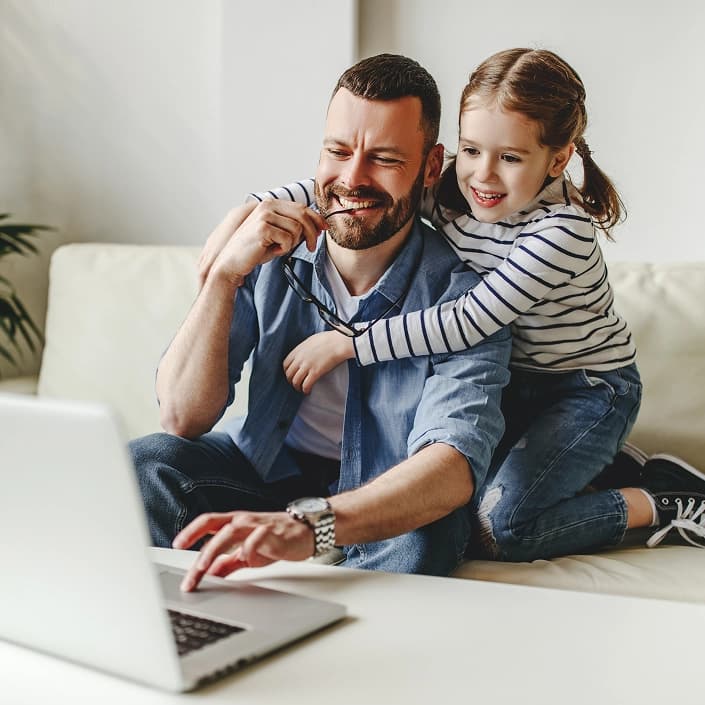 Smiling father using a laptop on the couch while his daughter playfully holds his glasses