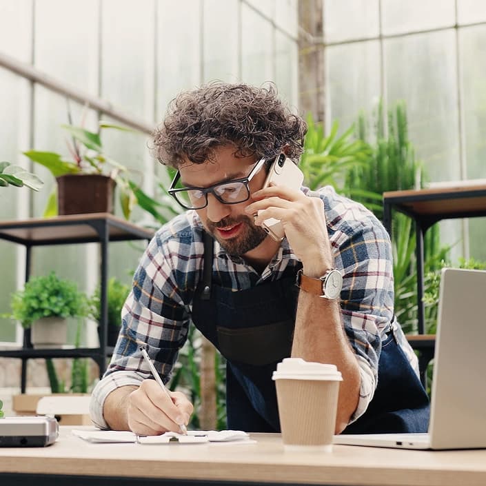 Small business owner talking on phone and taking notes at a desk surrounded by plants