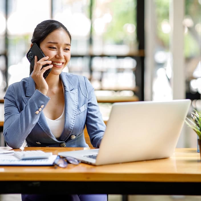 Smiling woman in a blue blazer talking on the phone while using a laptop for online banking.