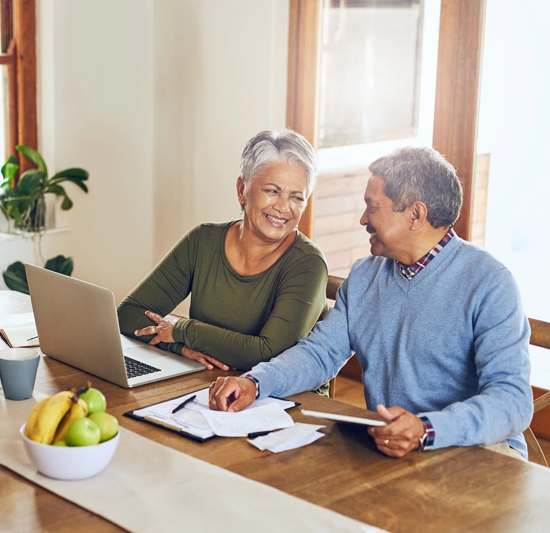 Happy couple smiling at each other while reviewing finances on a laptop at their kitchen table.