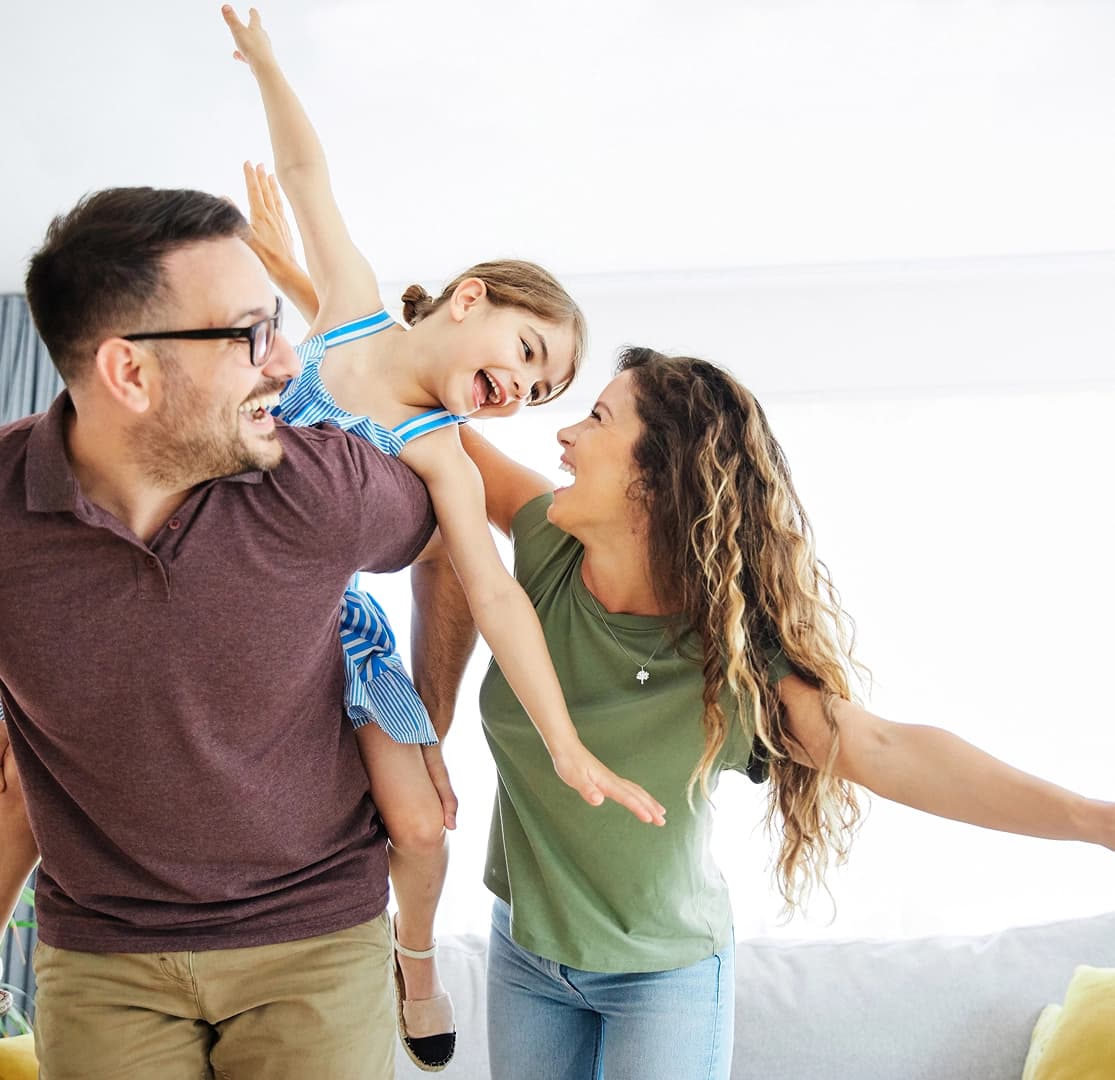 Parents playfully lifting their young daughter in a sunny living room