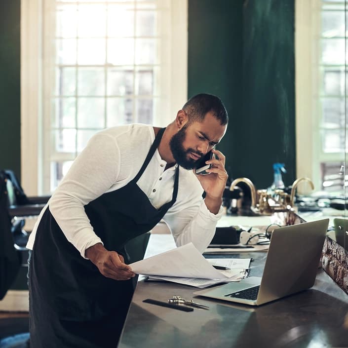 Small business owner reviewing paperwork while on a phone call near a laptop