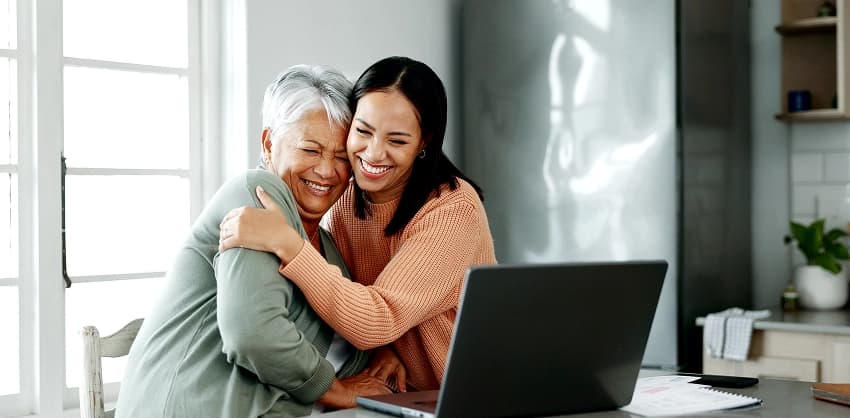 Two women smiling and hugging while reviewing online banking on a laptop at home