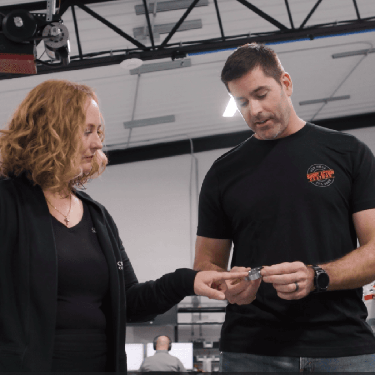 Two professionals examining a small component in a manufacturing facility, wearing Short Action Customs shirts