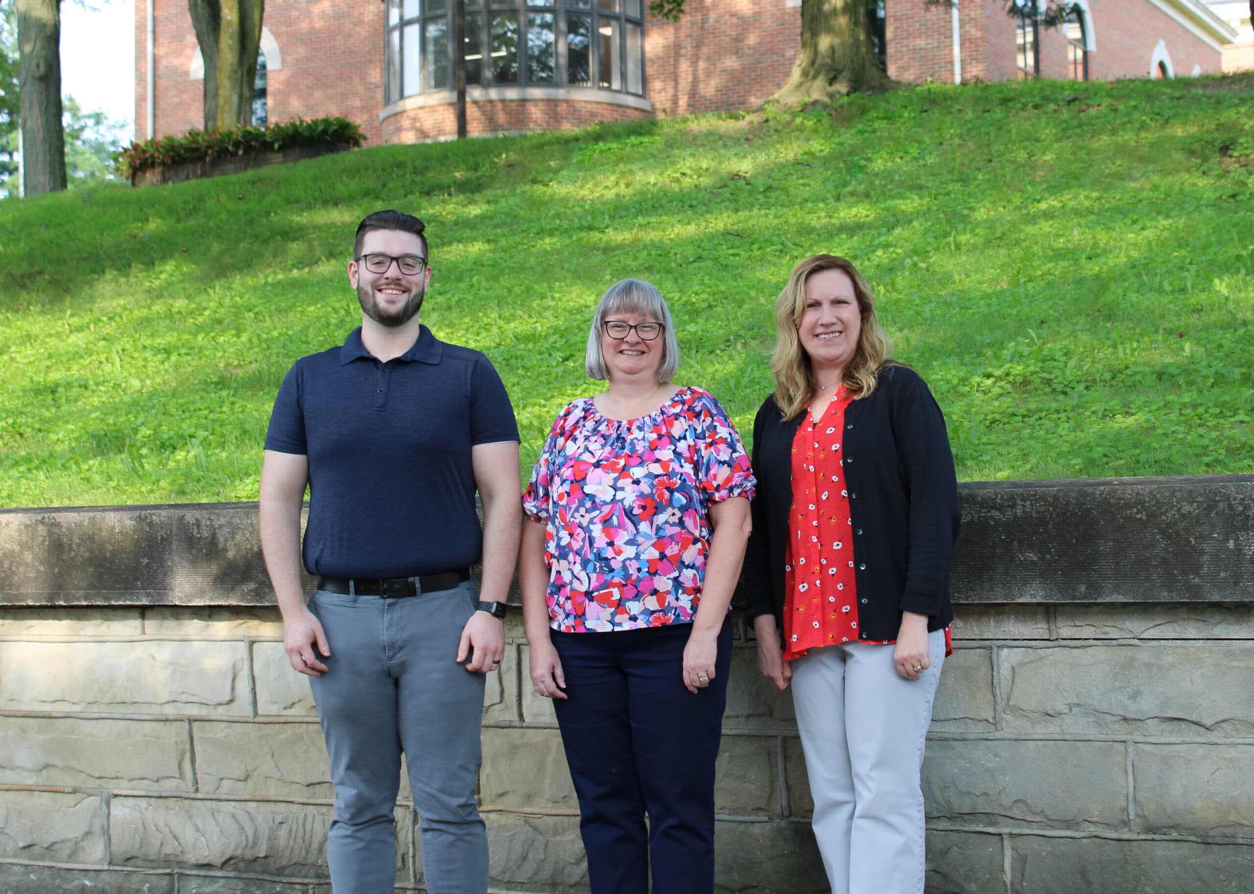Three Croghan Bank staff members smiling outdoors in front of a brick building on a green hillside.